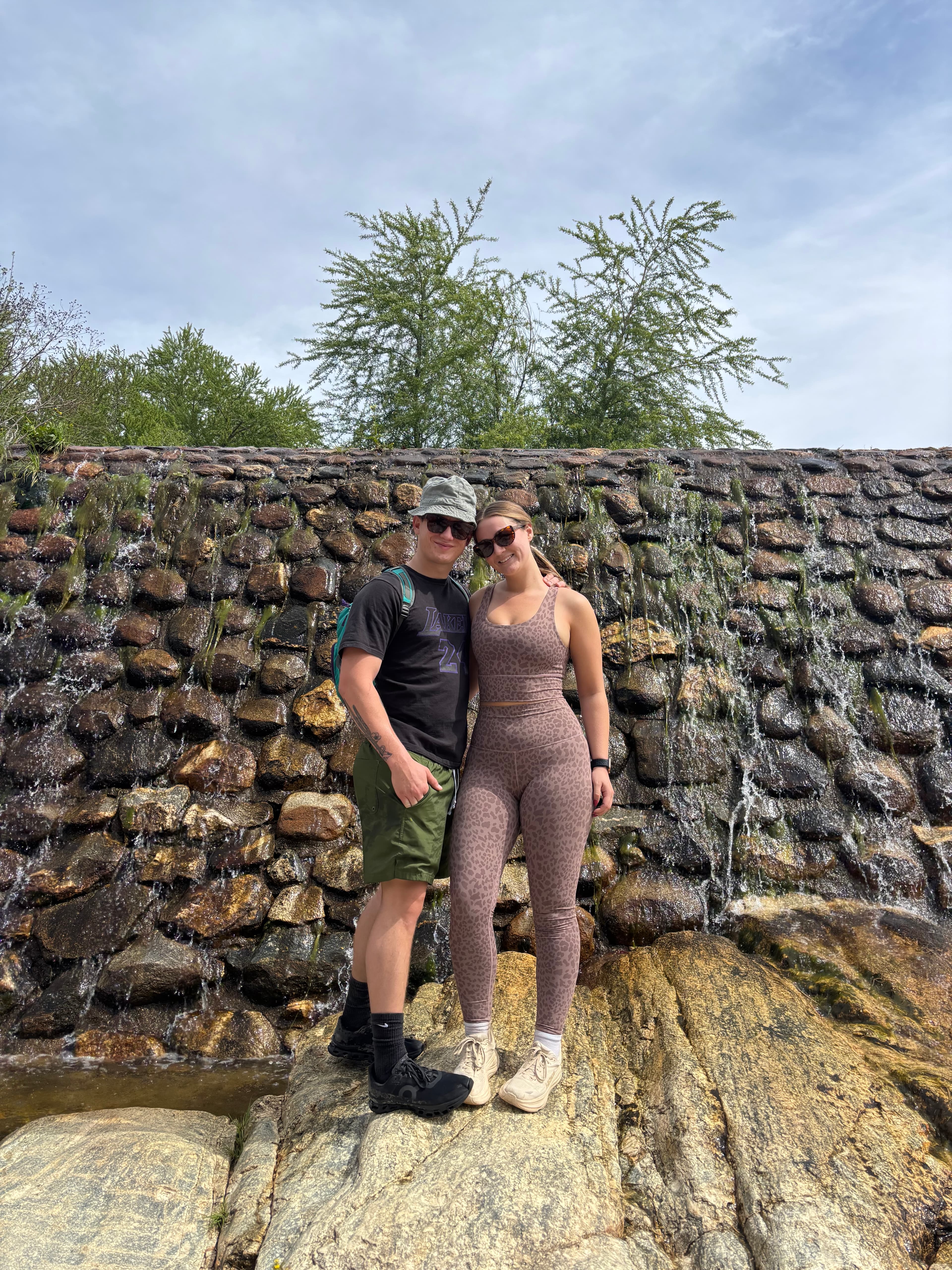 A couple in athletic gear poses before a stone wall with trickling water.